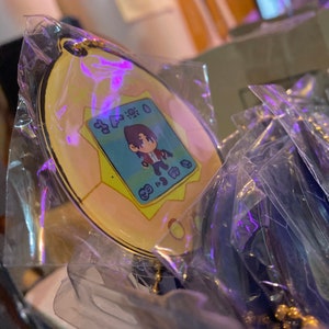 May include: A yellow and blue keychain featuring a cartoon character with brown hair and a red shirt. The keychain is shaped like a digital pet device with the text "8:30:00" on the screen.