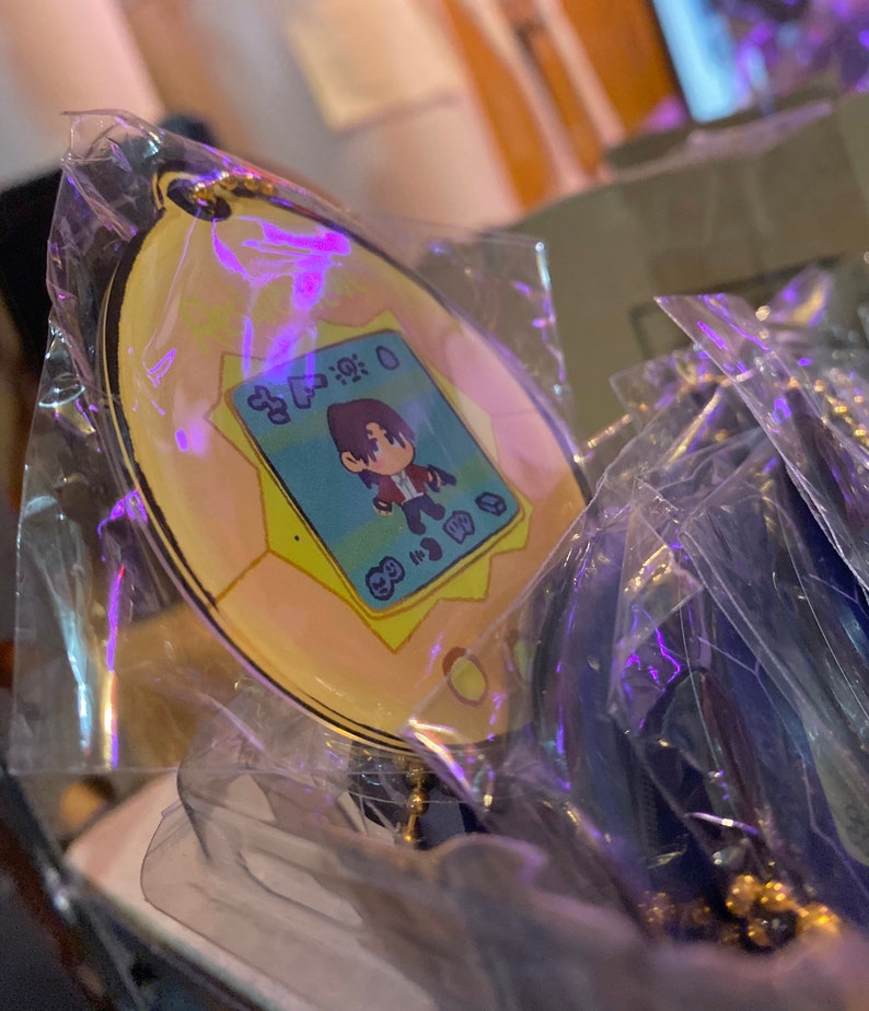 May include: A yellow and blue keychain featuring a cartoon character with brown hair and a red shirt. The keychain is shaped like a digital pet device with the text "8:30:00" on the screen.
