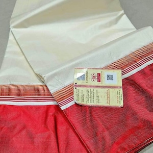 May include: Ivory-colored fabric with a red border and a tag attached. The fabric is folded, revealing a red section. The tag has text and a QR code. The fabric appears to be silk.
