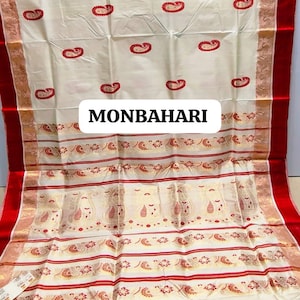 May include: Cream-colored textile with red paisley patterns and borders. The fabric features the text "MONBAHARI" in a white rectangular box. The edges are trimmed with red and gold accents, creating a decorative and elegant appearance.