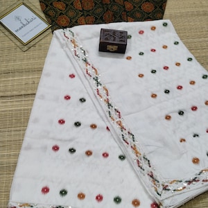 May include: White fabric with a colourful dot pattern and a decorative border. A dark brown blouse with a floral pattern is visible. A small wooden box and a framed sign are also present. The fabric is folded on a woven mat.