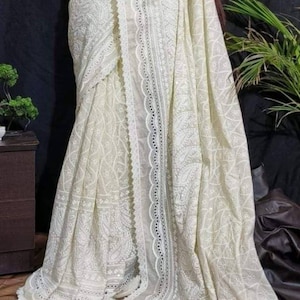 May include: Ivory-colored saree with intricate embroidered patterns and a delicate lace border. The saree drapes elegantly, showcasing detailed floral and geometric designs. A traditional garment, perfect for special occasions.