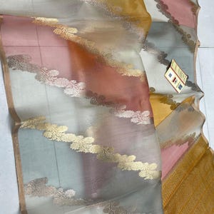 May include: A sheer, lightweight fabric drape featuring diagonal stripes in muted yellow, soft pink, and pale grey. The fabric is adorned with metallic gold floral-patterned borders running diagonally across the surface. Text reads KOR-IPR-135.