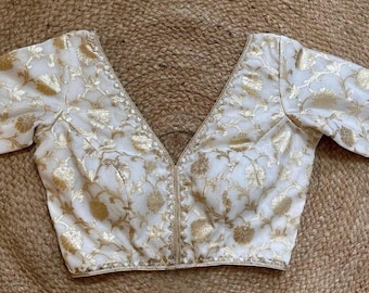 May include: Cream-coloured blouse with gold floral embroidery. The blouse has a deep V-neck and three-quarter sleeves. The edges of the neckline and sleeves are trimmed with gold detailing. The fabric has a subtle sheen, and the floral pattern is evenly distributed.