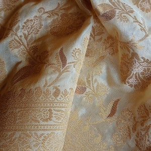 May include: A cream-colored fabric featuring an intricate gold metallic floral pattern woven throughout the material. The textile displays a dense botanical design with leaves and blossoms, showcasing a shimmering texture and a wide decorative border.