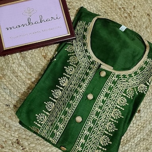 May include: A dark green tunic featuring gold-colored embroidery along the neckline and front placket, which includes four round buttons. A tag inside the collar displays the number 40. A framed sign nearby reads monbahari Tradition meets Modernity.