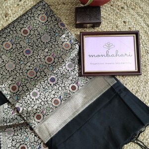 May include: A black saree featuring an intricate silver floral pattern with small red, blue, and green accents. The fabric has a wide silver border and a plain black end piece with tassels. A framed sign reads monbahari Tradition meets Modernity.