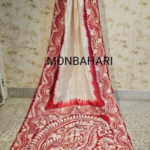 May include: Cream-coloured textile with a bold red border and intricate white floral patterns. The fabric is draped, showcasing the design and the text "MONBAHARI". The red border features a repeating floral motif, adding visual interest to the piece.