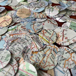 May include: A collection of circular paper cutouts, each featuring sections of detailed road maps. The maps display various colors and include place names like Charlotte and Colorado Springs. These map confetti pieces are scattered across a surface.