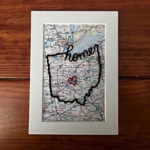 May include: Framed artwork featuring a map of Ohio with a black embroidered outline and the word "home". A red heart marks a location within the state. The artwork is set against a white mat and a wooden background.