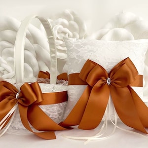 May include: White lace ring pillow with a large burnt orange satin bow and a small rhinestone in the center. A matching burnt orange satin bow decorates a white lace flower girl basket.