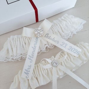 Peut inclure: Deux jarreti&egrave;res de mari&eacute;e blanches avec des n&oelig;uds en satin et des embellissements en strass. Une jarreti&egrave;re porte le texte "Mme Henderson" et l'autre porte le texte "Octobre 23, 2026".