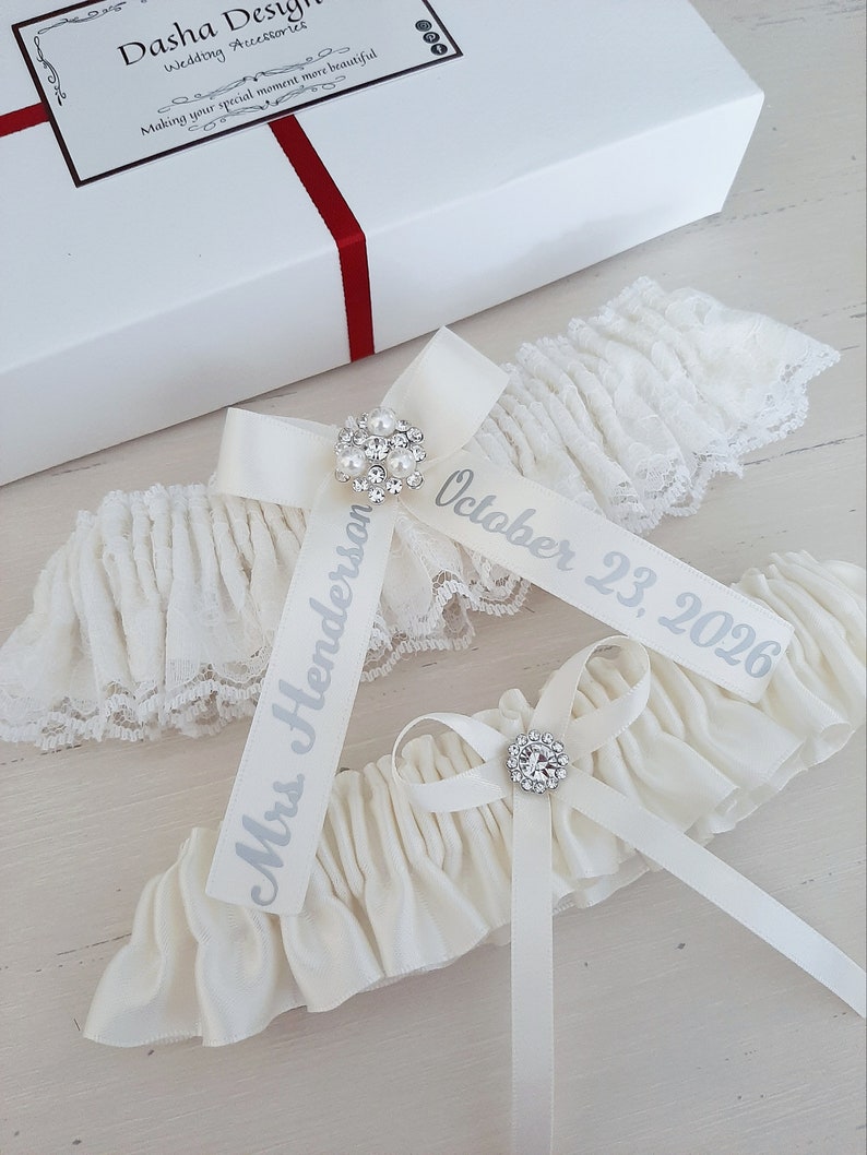 Peut inclure: Deux jarreti&egrave;res de mari&eacute;e blanches avec des n&oelig;uds en satin et des embellissements en strass. Une jarreti&egrave;re porte le texte "Mme Henderson" et l'autre porte le texte "Octobre 23, 2026".