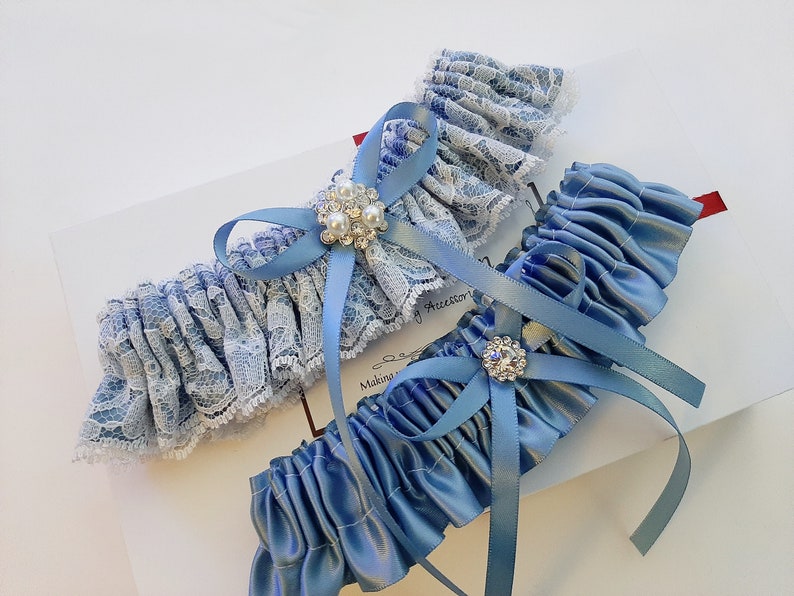 May include: Two blue garters with lace and satin. One garter has a white lace band with a blue bow and a rhinestone embellishment. The other garter is made of blue satin with a blue bow and a rhinestone embellishment.