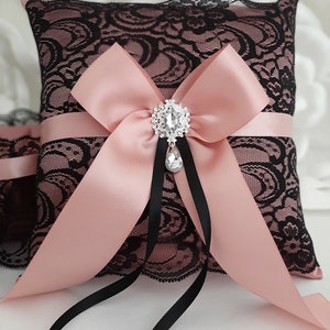 May include: A black lace ring pillow with a blush pink satin bow and a rhinestone brooch. The pillow has black satin ribbon ties.