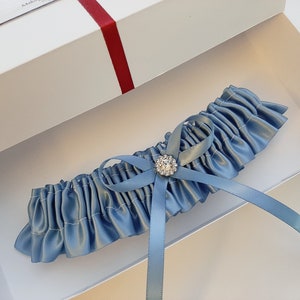 May include: A light blue satin garter with a small, clear rhinestone embellishment. The garter is in a white box with a red ribbon.
