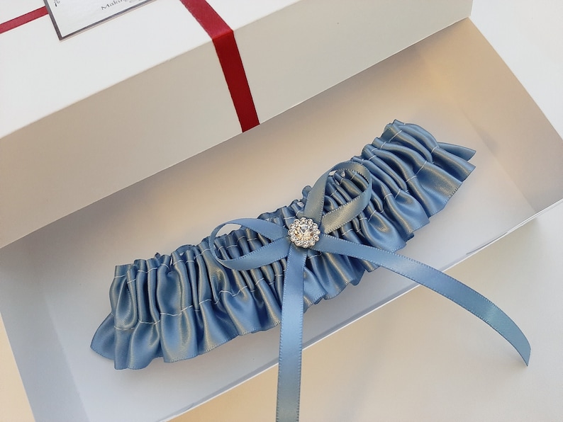 May include: A light blue satin garter with a small, clear rhinestone embellishment. The garter is in a white box with a red ribbon.
