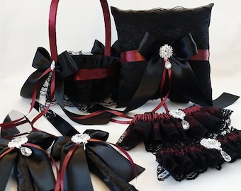 May include: A black lace wedding set with burgundy accents. The set includes a ring pillow, flower girl basket, garters, and two decorative wands. Each item features black satin bows, burgundy ribbons, and sparkling rhinestone embellishments.
