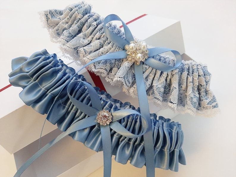 May include: Two blue garters with white lace and rhinestone embellishments. One garter is made of satin and the other is made of lace.