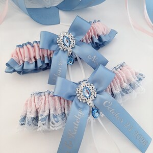 May include: Two wedding garters with blue bows and ribbons, featuring silver text and a jeweled centerpiece. The garters have pink and blue ruffled lace trim. One ribbon reads "Kevin & Katelyn October 23, 2026."