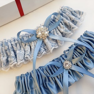 May include: Two blue garters with lace and satin. One garter has a blue bow with a rhinestone and pearl embellishment. The other garter has a blue bow with a rhinestone embellishment.