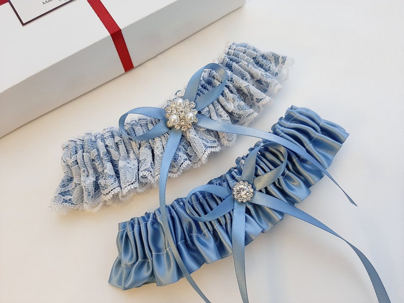 May include: Two blue garters with white lace and satin. One garter has a blue bow with a rhinestone and pearl embellishment. The other garter has a blue bow with a rhinestone embellishment.