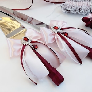 May include: A set of two cake cutting knives with pink and burgundy satin ribbon bows and jeweled accents. The knives are wrapped in burgundy satin ribbon and tied with a bow. The bows are accented with a silver and rhinestone brooch with a red jewel in the center.