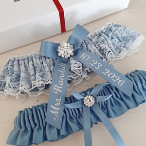 Peut inclure: Jarreti&egrave;re en satin bleu avec une bordure en dentelle blanche et un embellissement en strass. La jarreti&egrave;re est orn&eacute;e d'un ruban en satin blanc avec le texte "Mrs. Randall" et la date "10.23.2026" imprim&eacute;e dessus.