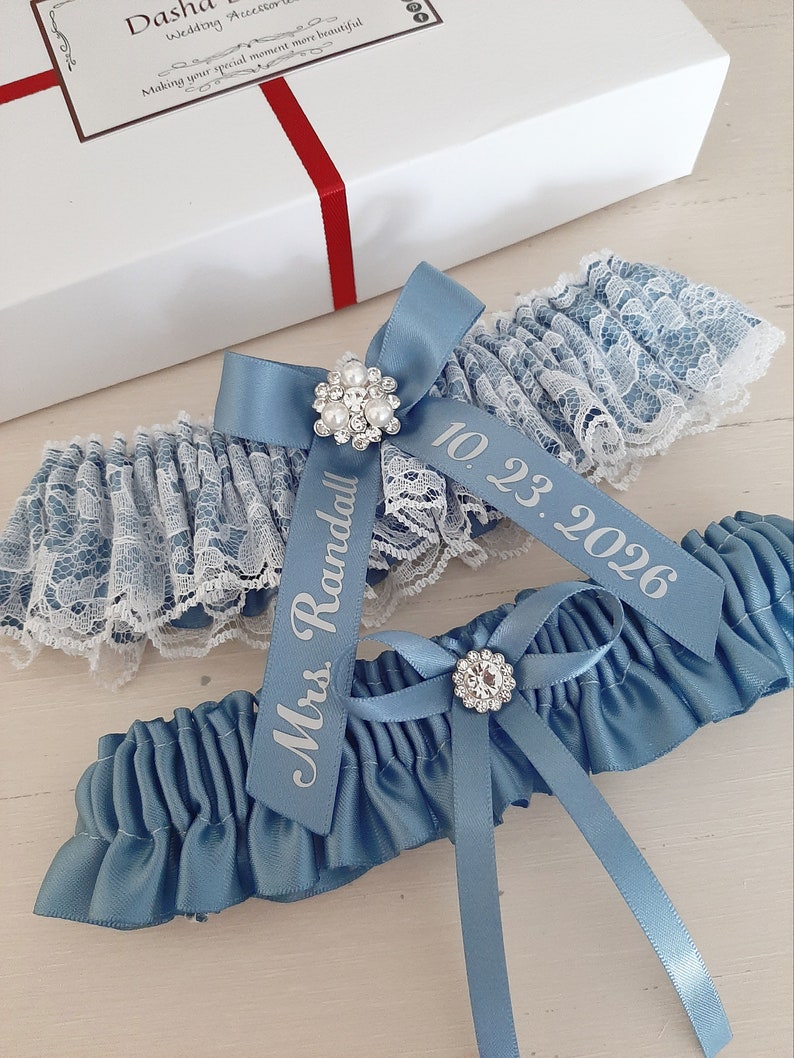 Peut inclure: Jarreti&egrave;re en satin bleu avec une bordure en dentelle blanche et un embellissement en strass. La jarreti&egrave;re est orn&eacute;e d'un ruban en satin blanc avec le texte "Mrs. Randall" et la date "10.23.2026" imprim&eacute;e dessus.
