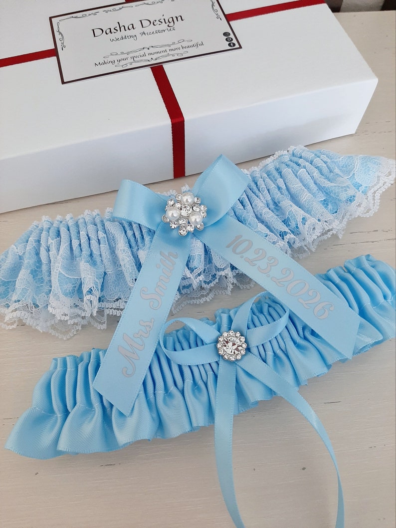 Peut inclure: Deux jarreti&egrave;res de mari&eacute;e bleu clair avec une bordure en dentelle blanche. La jarreti&egrave;re sup&eacute;rieure est orn&eacute;e d'un strass argent&eacute; et du texte "10.23.2026". La jarreti&egrave;re inf&eacute;rieure est orn&eacute;e d'un strass argent&eacute; et du texte "Mme Smith".