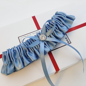 May include: A light blue satin garter with a small, clear rhinestone embellishment. The garter is tied with a bow and is displayed on a white box with red ribbon.