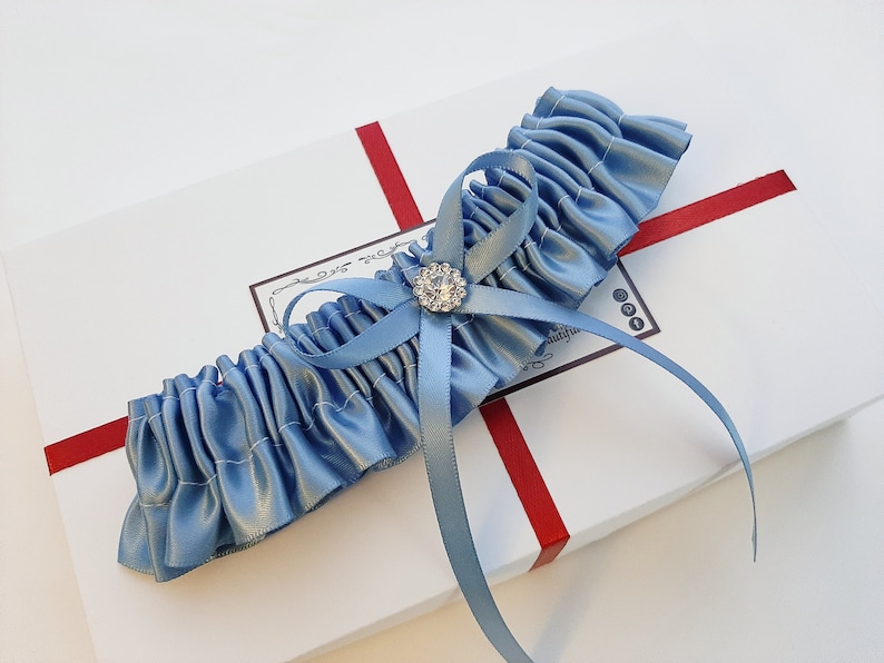 May include: A light blue satin garter with a small, clear rhinestone embellishment. The garter is tied with a bow and is displayed on a white box with red ribbon.