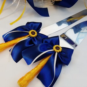 May include: A set of two cake cutting knives with yellow handles and blue ribbon bows. Each bow has a yellow sunflower accent.