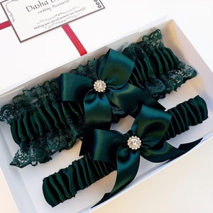 May include: Two green satin garters with lace trim and rhinestone embellishments in a white box.  The garters are tied with bows and are ready for a special occasion.