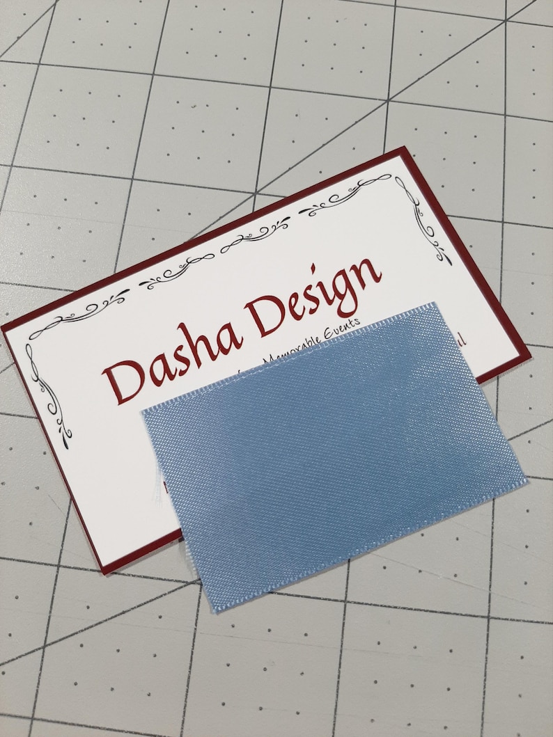 May include: A white business card with a red border and a decorative border design. The text "Dasha Design" is in red font. The text "Memorable Events" is in black font. A light blue ribbon is on top of the card.
