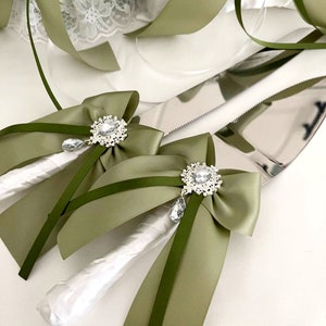 May include: Two white handled cake knives with olive green satin bows and rhinestone accents. The knives are on a white surface.