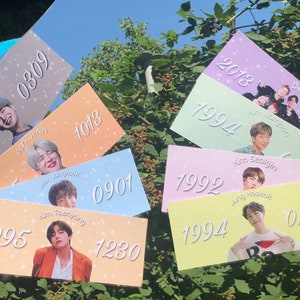 Puede incluir: Siete tarjetas coloridas con los nombres y los años de nacimiento de los miembros del grupo de K-pop BTS. Cada tarjeta presenta una foto de un miembro diferente, así como su nombre y su año de nacimiento en texto blanco.