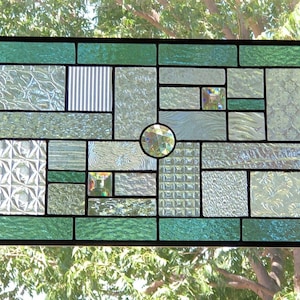 May include: A stained glass window with a geometric design featuring various shades of green and clear glass. The window is framed in black and has a small, round, clear glass piece in the center.