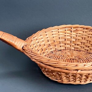 May include: A woven wicker basket with a long handle. The basket is oval-shaped and made of light brown wicker. The handle is also made of woven wicker and is attached to the side of the basket. The basket is likely used for serving or storage.