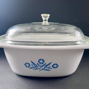 May include: A white rectangular casserole dish with rounded corners and a clear glass lid. The exterior of the dish features a blue floral pattern centered on the front. The lid includes a small, clear glass knob handle at the top center.