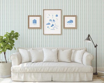 May include: A living room scene with a light-colored sofa, pillows, and framed art. The art features blue illustrations of buildings and a map. A green plant in a woven basket and a black floor lamp complete the decor. The wall has a vertical striped pattern.