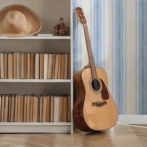 May include: Acoustic guitar with a natural wood finish leaning against a blue and white striped wall. A straw hat and a shelf of books are visible on a white bookshelf. The guitar has a dark brown pickguard.