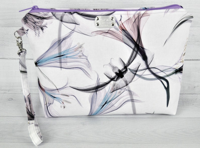 Large pouch in abstract cotton print with wrist strap attached on light background. White, black turquoise, plum and lavender flowers. Lavender zipper.