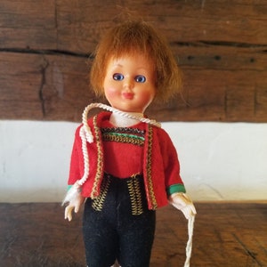 May include: A small doll dressed in a traditional Austrian costume, featuring red, black and gold colours. The doll has brown hair and blue eyes. The doll is holding a white string with a tag that reads "Made in Austria".