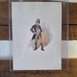Può includere: Una stampa di caricatura vintage incorniciata di un uomo con un cappello a cilindro, che tiene in mano una tazza. L'uomo indossa un cappotto nero, un gilet a righe e una sciarpa rossa. La stampa è in una custodia di plastica trasparente, esposta su una superficie di legno.