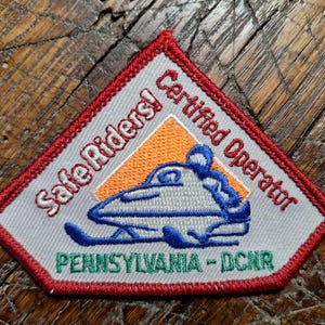 May include: Embroidered patch with a red border and the words "Safe Riders! Certified Operator." Features a blue and white snowmobile design with the text "PENNSYLVANIA - DCNR." The patch is on a wooden surface.