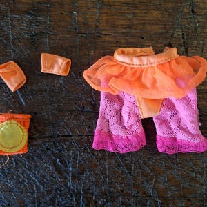 Op de afbeelding: Poppenkledingset met een tweedelige outfit met een geplooid topje en een broek met kanten patroon in oranje- en roze tinten. Inclusief bijpassende accessoires. De set is ontworpen voor een pop.
