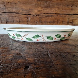 May include: An oval-shaped ceramic baking dish with a cream-colored base and a dark green rim. The exterior features a repeating pattern of green holly leaves and small red berries connected by thin brown stems encircling the entire vessel.