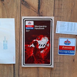 Puede incluir: Un manual de membresía del Amoco Motor Club rojo, blanco y azul con una ilustración vintage de un automóvil siendo remolcado. El manual incluye una lista de beneficios de membresía y una calcomanía con el logotipo del Amoco Motor Club.