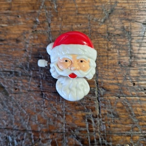 May include: A wind-up Santa Claus head figurine featuring a bright red hat with white trim, a peach-toned face, dark eyes, a white moustache, and a full white beard. A small white plastic winding key protrudes from the left side of the head.